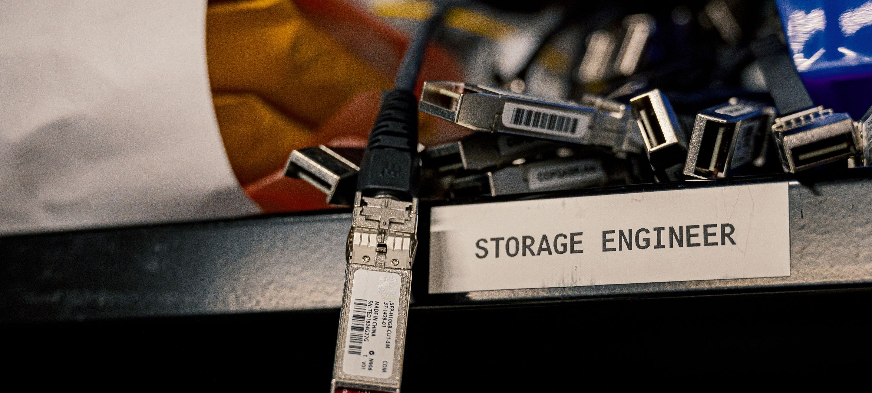 Spare optical transceivers and cables on a storeroom shelf beside a nameplate reading Storage Engineer