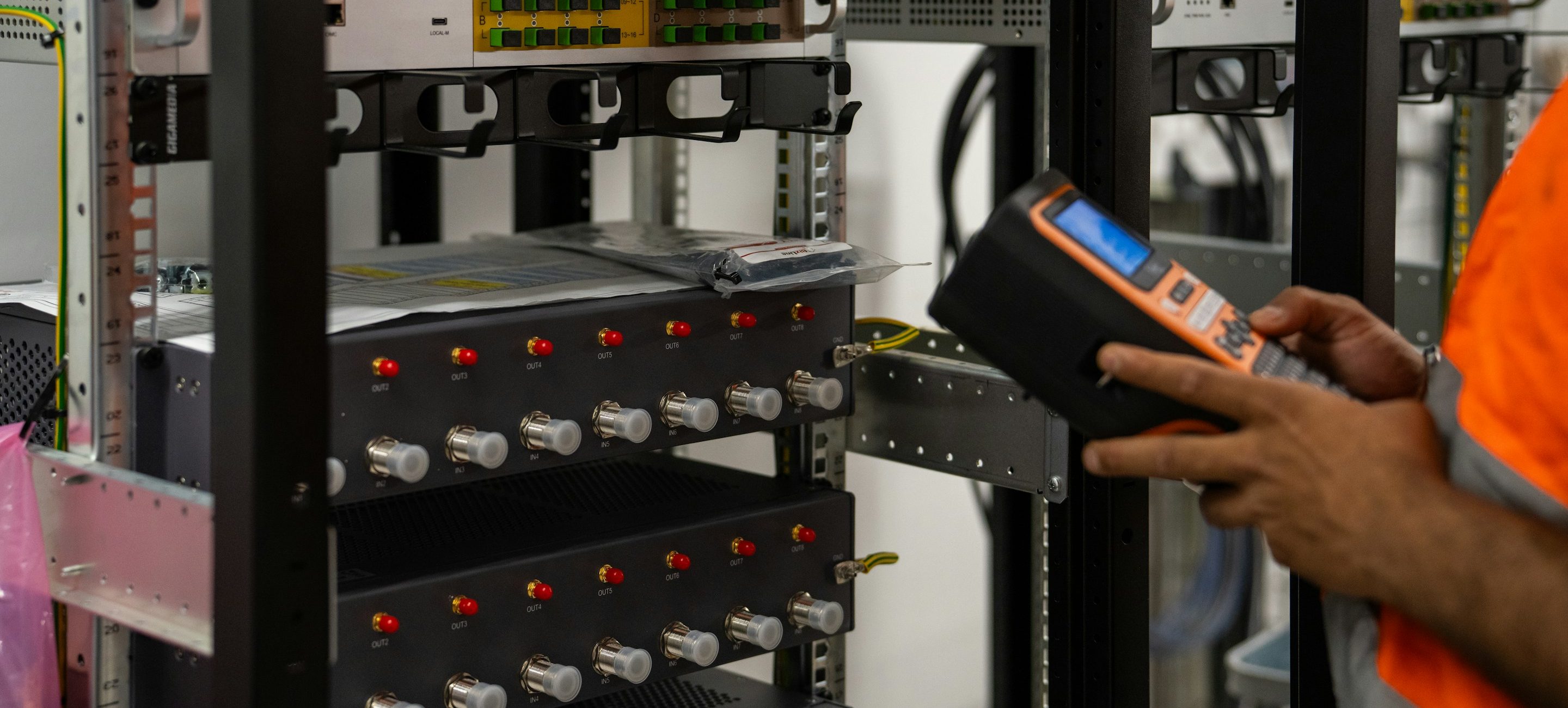A technician in a high-visibility vest reading a rack of modules with a handheld test instrument
