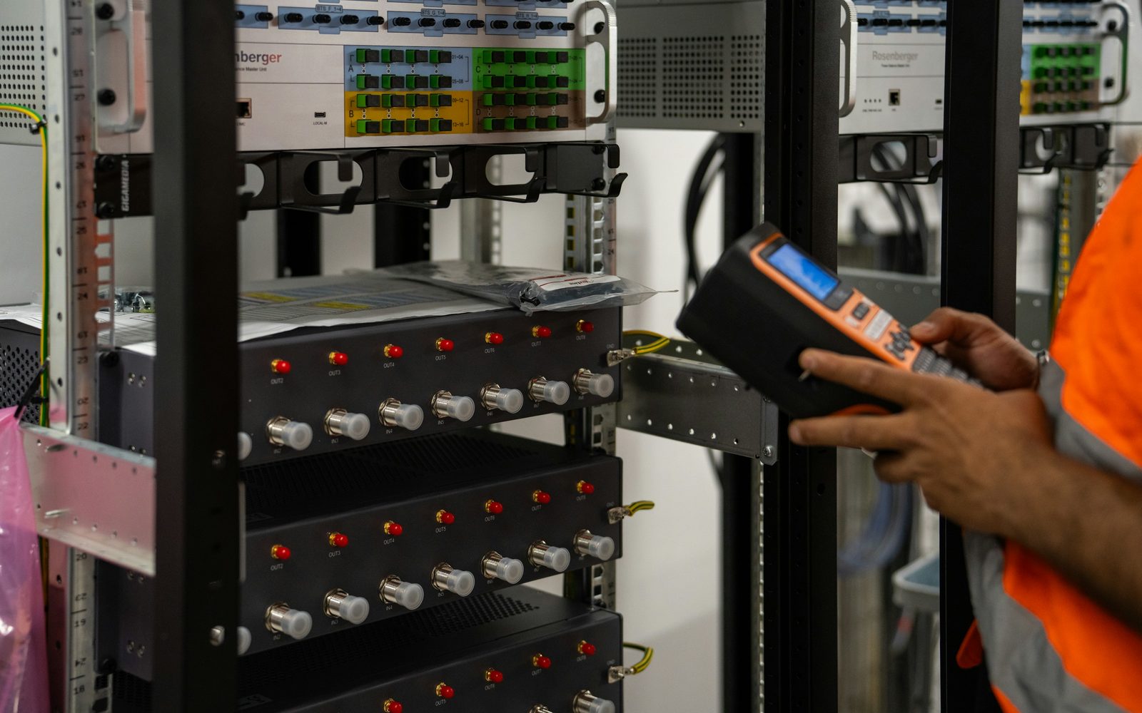 A site engineer holding a handheld tester in front of a cabinet of patch panels and rack-mounted units