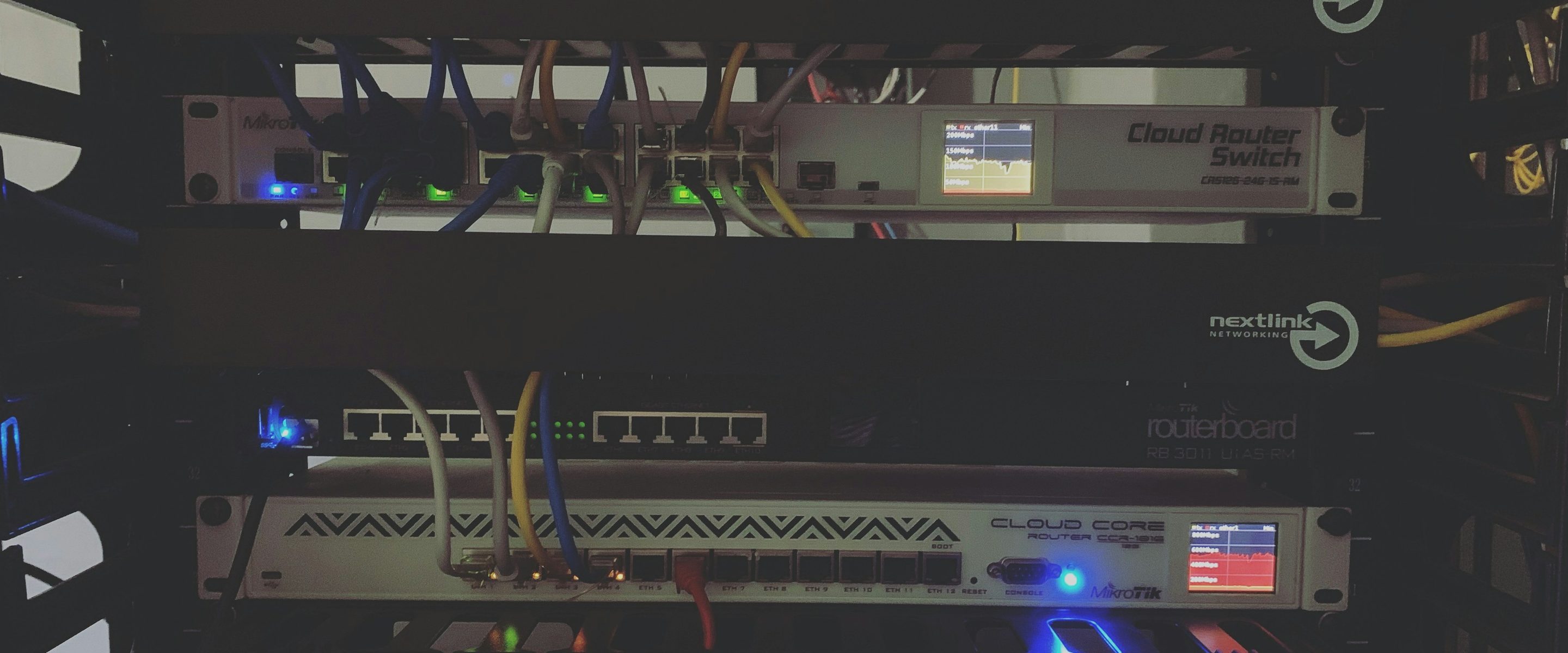 Routers and a switch in a dimly lit rack, indicator lights and small status screens showing